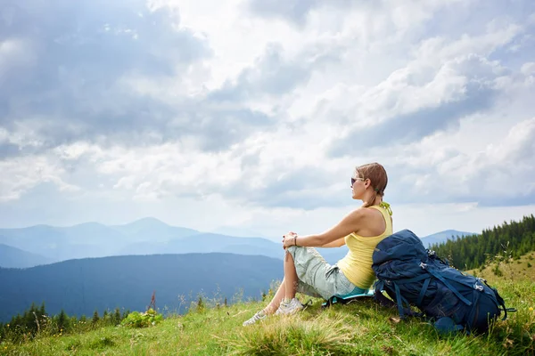 Yan görünüm ile sırt çantası, çimenli tepe üzerinde dinlenme çekici kadın hiker güneş gözlüğü takıyor. Kadın turist Karpat Dağları yaz bulutlu gün zevk. Açık hava etkinliği, turizm kavramı