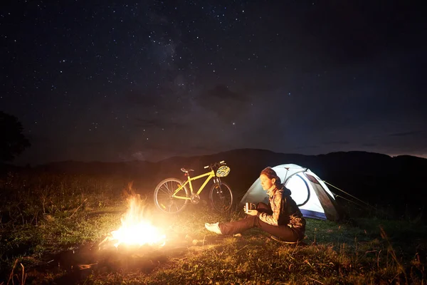 Genç kadın yolcu gece kamp yanan kamp ateşi, ışıklı turist çadır, güzel bir akşam gökyüzü yıldızlarla dolu altında dağ bisikleti yakınındaki, zevk. Açık kavramı, etkinlik ve Turizm
