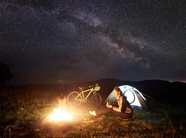 Genç kadın turist gece kamp ateşi, ışıklı turist çadır, dağ bisikleti inanılmaz güzel akşam gökyüzünde yıldızlar ve Samanyolu dolu altında yanan yakınındaki kampa adlı bir dinlenme. Turizm kavramı