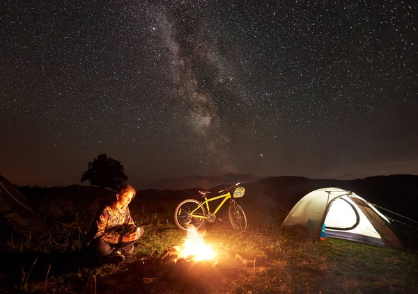 Genç kadın turist kamp ateşi, ışıklı turist çadır, dağ bisikleti inanılmaz güzel akşam gökyüzünde yıldızlar ve Samanyolu dolu altında yanan yakınındaki gece kamp dinlenme. Turizm kavramı