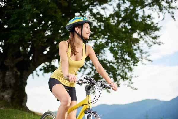 Miğfer, dağlarda yaz günü zevk çekici mutlu kadın bisikletçi büyük ağacın altında sarı dağ bisiklet sürme. Açık spor aktivitesi, yaşam kavramı