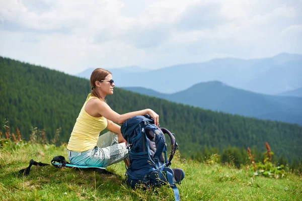 Güneş gözlüğü, Karpat Dağları yaz bulutlu gün zevk giyiyor oturma ve sırt çantası ile çimenli tepe üzerinde dinlenme çekici kadın backpacker yan görünüm. Açık hava etkinliği, turizm kavramı
