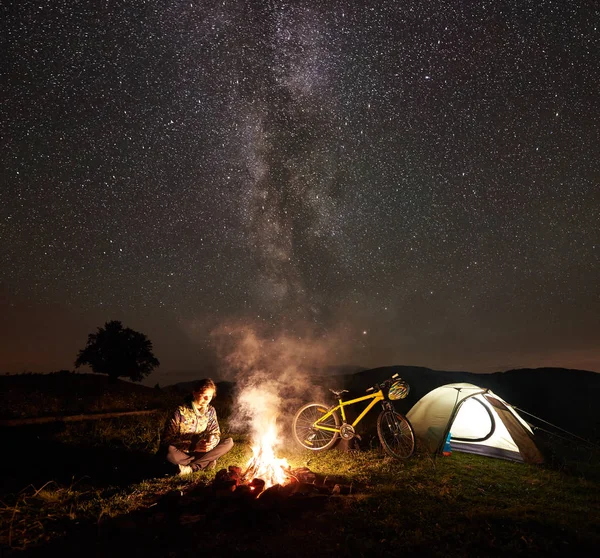 Genç kadın bisikletçi gece kamp kamp ateşi, ışıklı turist çadır, dağ bisiklet inanılmaz güzel akşam gökyüzünde yıldızlar ve Samanyolu dolu altında yanan yakınındaki zevk. Astrophotography