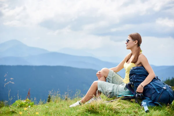 Yan görünüm sırt çantası ve trekking, sopa ile çimenli tepe üzerinde dinlenme çekici kadın hiker güneş gözlüğü, dağlarda yaz bulutlu gün zevk takıyor. Açık hava etkinliği, turizm kavramı
