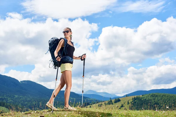 Young happy woman backpacker hiking mountain trail, walking on grassy hill, wearing backpack and sunglasses, using trekking sticks, enjoying summer sunny day in the mountains. Tourism concept