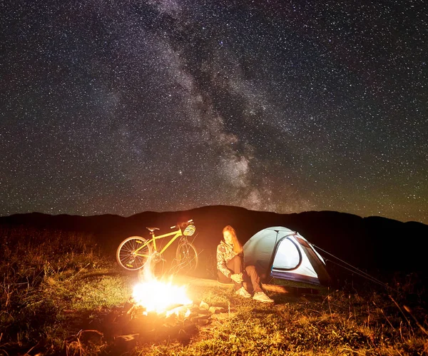 Genç kadın turist gece kamp ateşi, ışıklı turist çadır, dağ bisikleti inanılmaz güzel akşam gökyüzünde yıldızlar ve Samanyolu dolu altında yanan yakınındaki kampa adlı bir dinlenme. Turizm kavramı