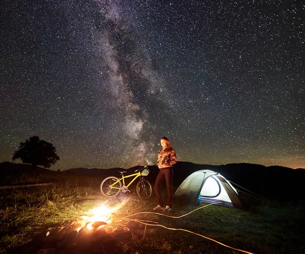 Genç kadın bisikletçi gece kamp ateşi, ışıklı turist çadır, dağ bisiklet inanılmaz güzel akşam gökyüzünde yıldızlar ve Samanyolu dolu altında yanan yakınındaki kampa adlı bir dinlenme. Astrophotography