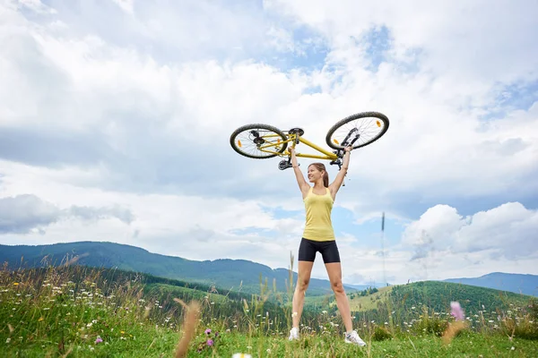 Attractive happy girl cyclist holding yellow mountain bike above a head, enjoying summer day in the mountains. Outdoor sport activity, lifestyle concept