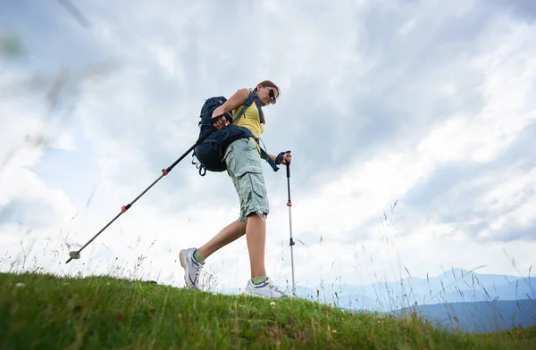 Low angle view of attractive woman tourist hiking in Carpathian mountain trail, walking on grassy hill, wearing backpack and sunglasses, using trekking sticks, enjoying summer day. Lifestyle concept