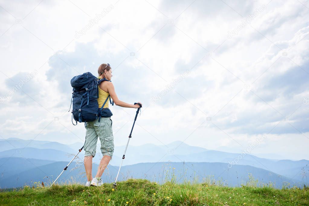 Back view of athlete woman backpacker hiking mountain trail, walking on grassy hill, wearing backpack, using trekking sticks, enjoying summer day in the mountains. Outdoor activity, lifestyle concept