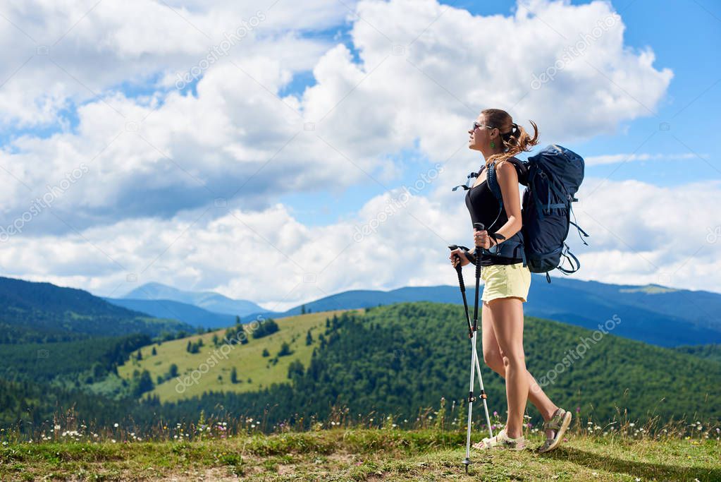 Attractive woman tourist hiking mountain trail, walking on grassy hill, wearing backpack and sunglasses, using trekking sticks, enjoying summer sunny day in the Carpathian mountains. Tourism concept