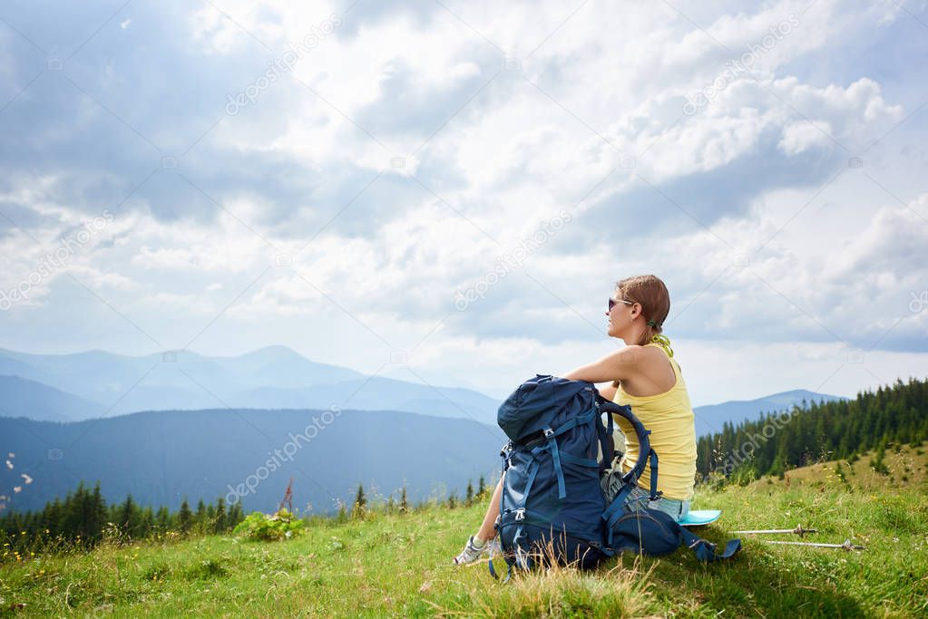 Back view of attractive woman tourist resting on grassy hill with backpack and trekking sticks. Female backpacker enjoying summer cloudy day in the mountains. Outdoor activity, tourism concept