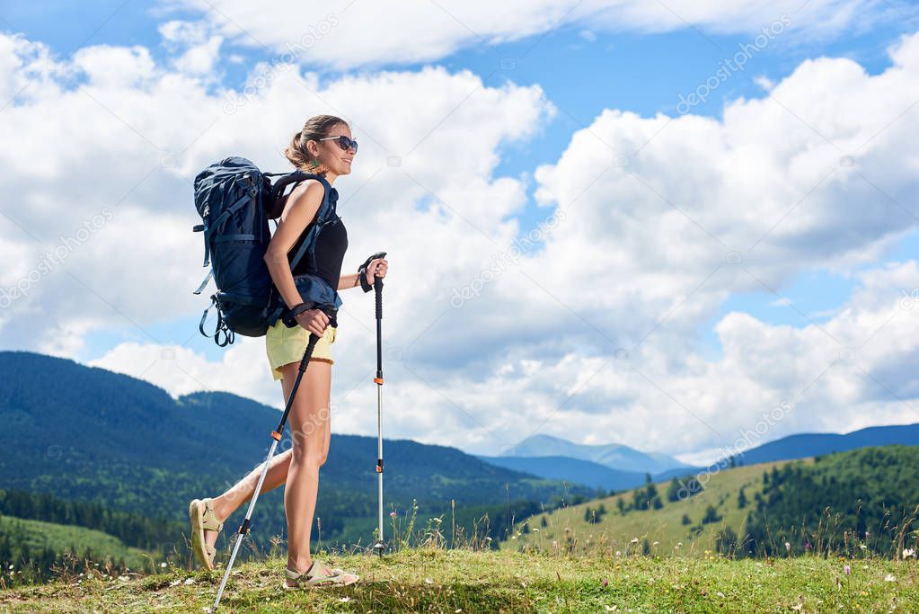 Attractive smiling woman tourist hiking mountain trail, walking on grassy hill, wearing backpack, using trekking sticks, enjoying summer sunny day in the Carpathian mountains. Tourism concept