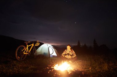 Genç kadın bisikletçi gece kamp yanan kamp ateşi, ışıklı turist çadır, güzel bir akşam gökyüzü yıldızlarla dolu altında dağ bisikleti yakınındaki, zevk. Açık kavramı, etkinlik ve Turizm