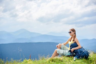 Yan görünüm ile sırt çantası, çimenli tepe üzerinde oturan çekici kadın backpacker güneş gözlüğü, Karpat Dağları yaz bulutlu gün zevk takıyor. Açık hava etkinliği, turizm kavramı
