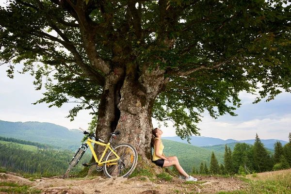 Attractive happy woman cyclist resting near yellow mountain bicycle under big tree, enjoying summer day in the mountains. Outdoor sport activity, lifestyle concept