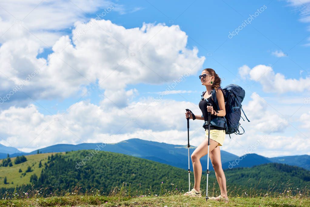 Young woman backpacker hiking mountain trail, walking on grassy hill, wearing backpack and sunglasses, using trekking sticks, enjoying summer sunny day in the mountains. Tourism concept