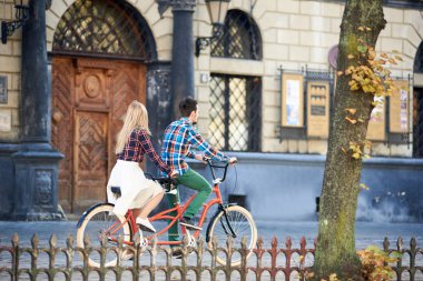 Parlak güneşli bulanık Antik Bina zemin üzerine genç turist çift, sakallı adam ve beyaz etek sürme iki kişilik bisiklet döşeli şehir kaldırım boyunca uzun saçlı sarışın kadın görünümünü geri.