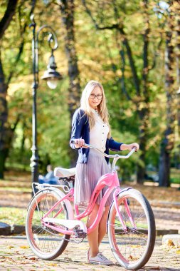 Young happy attractive blond long-haired woman in glasses, skirt and blouse standing with pink lady bicycle on sunny paved park alley on blurred golden yellow green bokeh foliage background.