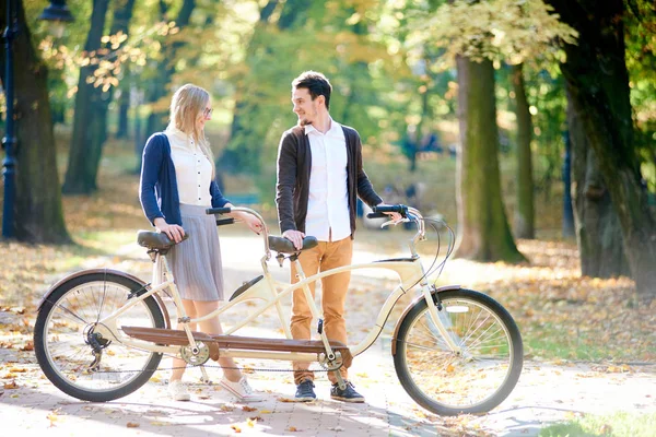 Young happy smiling couple, handsome bearded man and attractive blond woman standing together at tandem double bike on sunny paved path covered with golden leaves on blurred tall trees background.