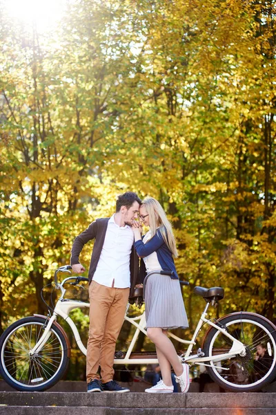 年轻幸福浪漫情侣在爱情 胡子的家伙和迷人的女孩站在一起在秋季公园或森林中的双联自行车在阳光明媚的金黄叶子散景背景 — 图库照片