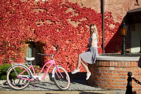 Attractive long-haired smiling blond girl in short skirt and blouse l sitting on low brick wall at pink lady bicycle on bright warm sunny day on background of house overgrown with beautiful red ivy.