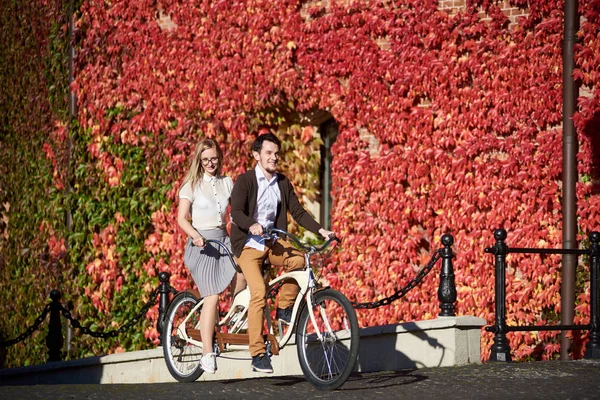 若い幸せ観光客カップル ハンサムなあごひげを生やしたと完全に赤いツタの生い茂った建物の背景に明るい晴れた日にかなり長い髪のブロンド女性サイクリング タンデム自転車の葉 — ストック写真