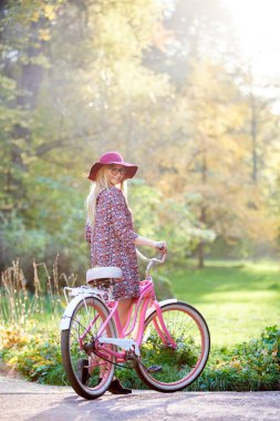 İnce sarışın moda uzun saçlı çekici kız gözlük, kısa elbise ve Bayan bisikletle döşeli güneşli yaz park sokak güzel Sisli Yeşil ve altın ağaçlar arka plan üzerine açık havada, pembe şapka.