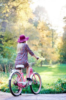 Sonbaharda güzel yeşil ve altın ağaçlar arka plan üzerinde ince şık uzun saçlı çekici sarışın kısa elbise ve Bayan Bisiklet döşeli park sokak üzerinde kırmızı şapka, arkadan görünüm.