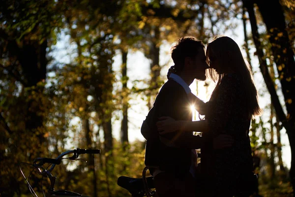 点燃了夕阳轮廓的浪漫情侣 年轻男子和迷人的女孩密切在一起在黑暗的秋季公园或森林在昏暗的金色散景背景下的串联自行车户外 — 图库照片
