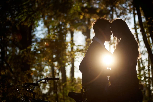 点燃了设置太阳剖面剪影浪漫情侣 年轻男子和迷人的女人紧密在一起在黑暗的秋季森林在昏暗的金色散景背景下的串联自行车户外 — 图库照片