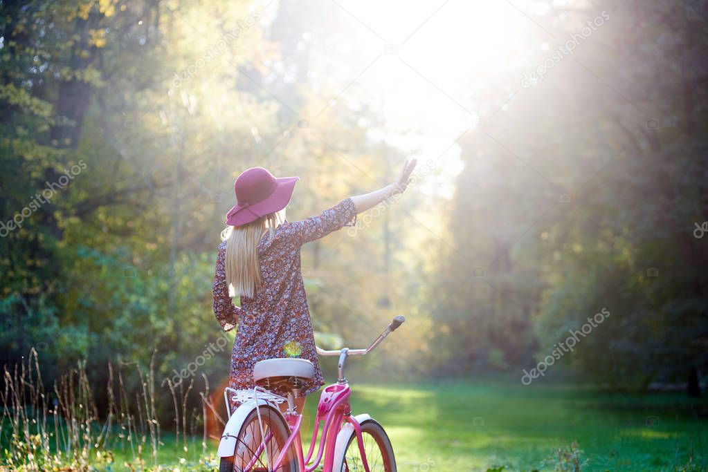 Back view of slim blond fashionable long-haired attractive girl in short dress and pink hat with raised arm at lady bicycle on paved summer park alley, enjoying beautiful evening sunset
