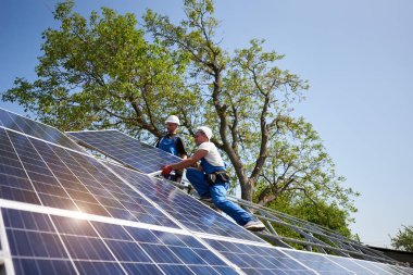 İki işçi yeşil ağaç ve mavi gökyüzü arka plan ağır güneş Fotoğraf volta panelini takma çelik platformda koruyucu kask içinde. Dış güneş paneli sistemi kurulması, tehlikeli bir iş kavramı.