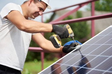 Arkadan görünüşü güneş Fotoğraf volta paneli parlak güneşli yaz gününde Elektrikli tornavida kullanarak metal platformu bağlanma kask içinde Genç teknisyen. Tek başına güneş paneli sistemi kurulması.