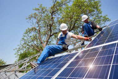 İki teknisyen parlak mavi gökyüzü ve yeşil ağaç üreten güneş paneli sistemi yüklemesi üzerinde çalışma karşı yüksek metal platformda. Yenilenebilir ucuz yeşil enerji üretim kavramı.