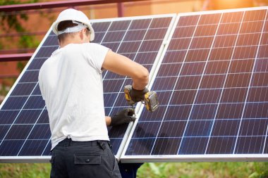 Arkadan görünüşü Genç teknisyen güneş Fotoğraf volta paneli metal platformu üzerinde parlak güneşli yaz gün açık havada Elektrikli tornavida kullanarak bağlanma. Alternatif yenilenebilir enerji üretimi kavramı.