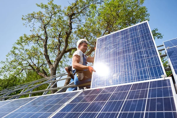 青い空と緑の木の背景に重い太陽太陽光発電パネルを調整する金属のプラットフォームの上に座って若い技術者 スタンドアロンの太陽電池パネル システムのインストールとプロフェッショナ リズムの概念 — ストック写真