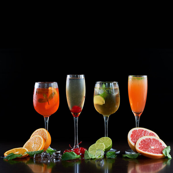 Composition with four different shapes crystal cocktail glasses with cold drinks, orange, lime and grapefruit slices, melting ice cubes, mint leaves and red cherries on black background. Ratio 1:1