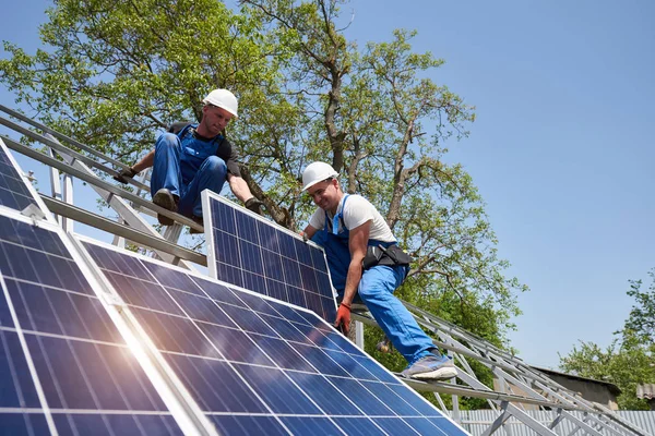 Two young technicians mounting heavy solar photo voltaic panel on tall steel platform on green tree background. Exterior solar panel voltaic system installation, dangerous job concept.