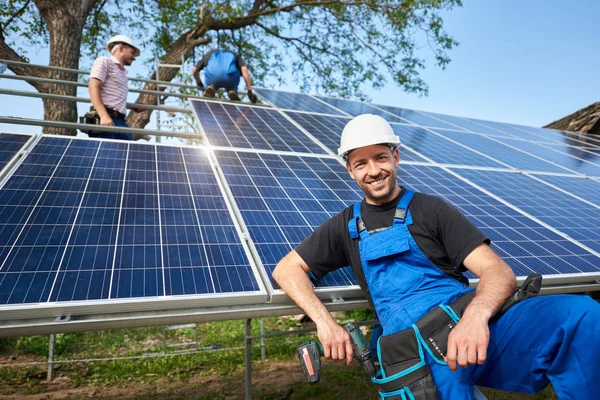 在未完成的高外部太阳能电池板光电系统前面的微笑技术员的画像在高平台上的工作者团队 — 图库照片