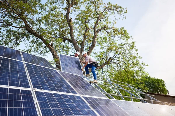 Two technicians mounting heavy solar photo voltaic panel on tall steel platform on green tree and blue sky background. Exterior solar panel voltaic system installation, dangerous job concept.