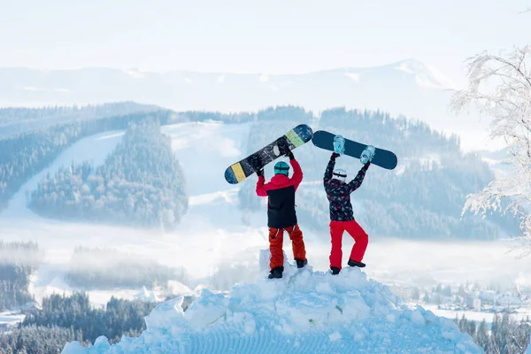 Two people standing with their backs on a snowdrift and raise their snowboards up against a white haze of snow-capped mountains and forests at winter ski resort.