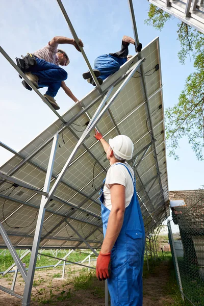 View from inside of stand-alone exterior solar voltaic system installing in rural countryside. Engineer technician pointing team of workers on high steel platform on bright clear blue sky background.