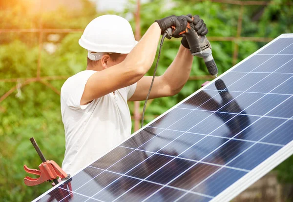 Profile of professional worker in helmet connecting solar photo voltaic panel to metal platform using electrical screwdriver outdoors on bright sunny day on blurred green rural landscape background.