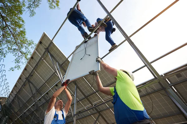 Bottom view from inside of stand-alone exterior solar panel system with team of engineers working on high steel platform on bright clear blue summer sky background. Green energy production concept.