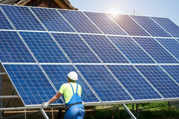 安全ヘルメットと鋼プラットフォームにインストールされている高外観ソーラー パネル太陽光発電システムに傾いて青いオーバー オールの労働者の背面します 生態学的なグリーン エネルギー生産のコンセプト ストック写真