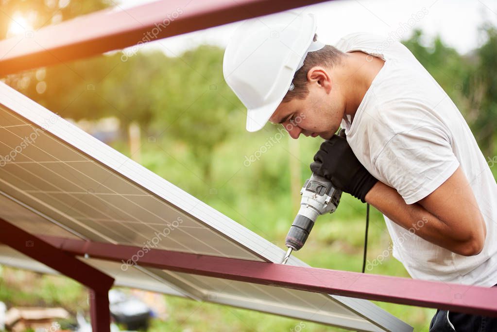 Profile of professional technician working with screwdriver connecting solar photo voltaic panel to metal platform on green summer background. Stand-alone exterior solar panel system installation.