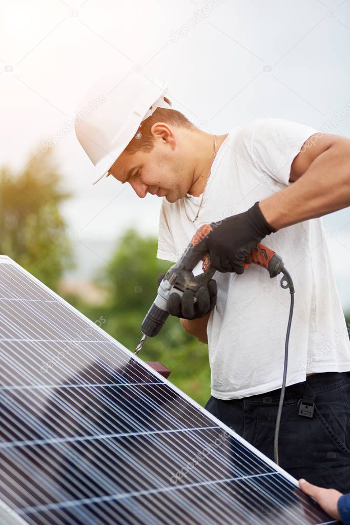 Close-up of professional worker in helmet and protective gloves connecting solar photo voltaic panel to platform using electrical screwdriver on bright sunny sky and green trees background.