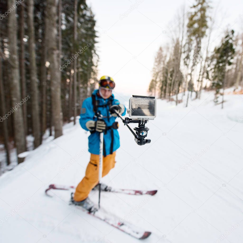 Skier man taking selfie with stick over forest on the winter resort. Focus on his camera, blurred background