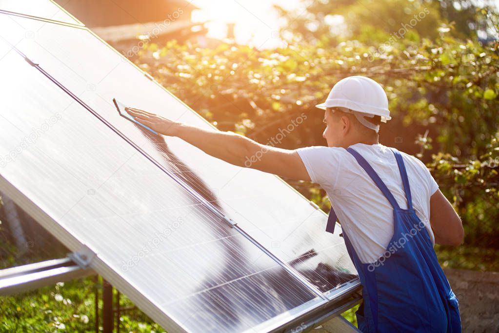 Profile of worker in white helmet and blue overalls wiping clean big square shiny solar panel on sunny summer bright green foliage bokeh background. Renewable energy generation concept.